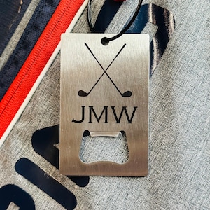 May include: A silver metal bottle opener with a golf club design and the initials "JMW" engraved on it. The bottle opener has a built-in bottle opener.