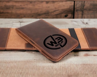 Puede incluir: Un porta tarjetas de puntuación de golf de cuero marrón con un logotipo negro que dice "WB GOLF" en un círculo.