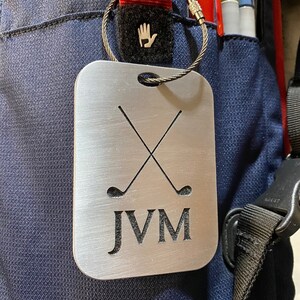 May include: A silver metal golf bag tag with a brushed finish. The tag features two crossed golf clubs and the initials "JVM".