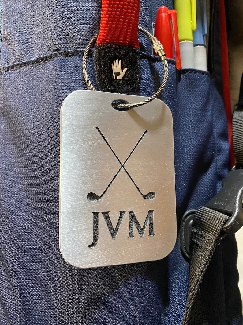 May include: A silver metal golf bag tag with a brushed finish. The tag features two crossed golf clubs and the initials "JVM".