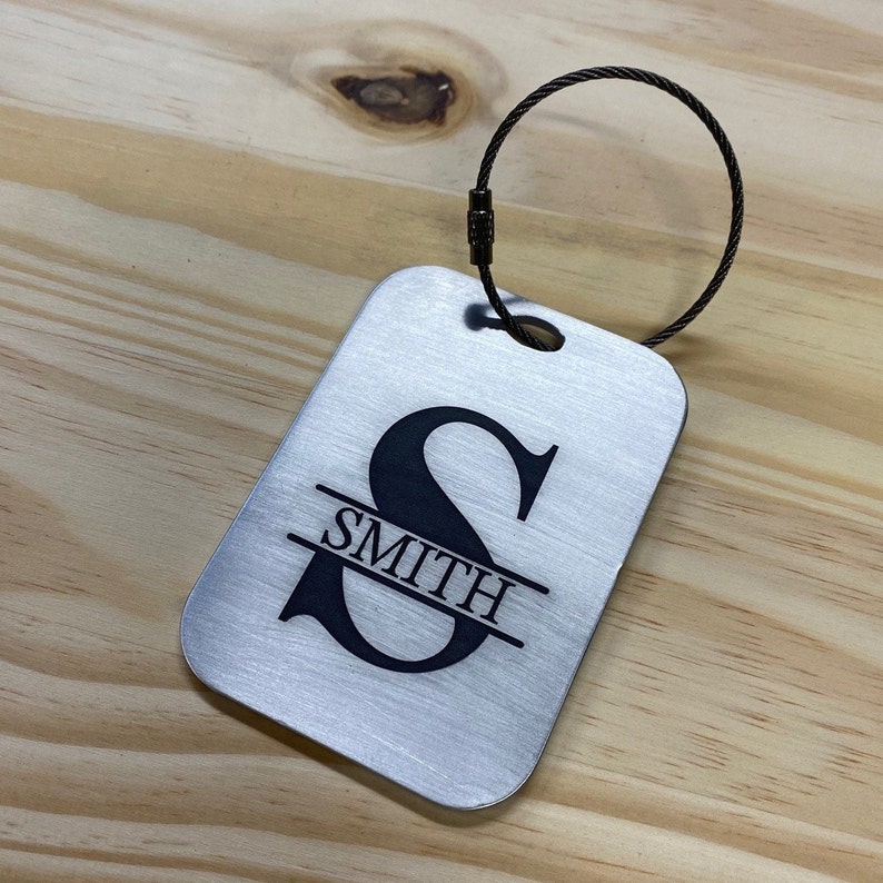 May include: Silver luggage tag with a black cable loop. The tag has a black engraved letter "S" with the name "SMITH" centered below the letter.