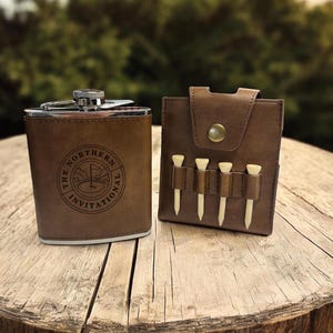 May include: A brown leather flask with a silver top and a circular engraved design that says "The Northern Invitational". The flask is next to a brown leather golf tee holder with four white golf tees.