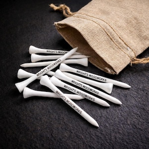 May include: White golf tees with the text "WRIGHT'S WOODWORKS" printed in black, scattered on a dark surface. A tan burlap bag is in the background. The tees are made of wood and are approximately 3 inches long.