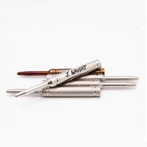 May include: A collection of metallic pens with a pointed tip and a cylindrical body featuring horizontal grooves. One silver-toned pen is positioned on top, clearly engraved with the name J. WRIGHT in black lettering, while others lie beneath.