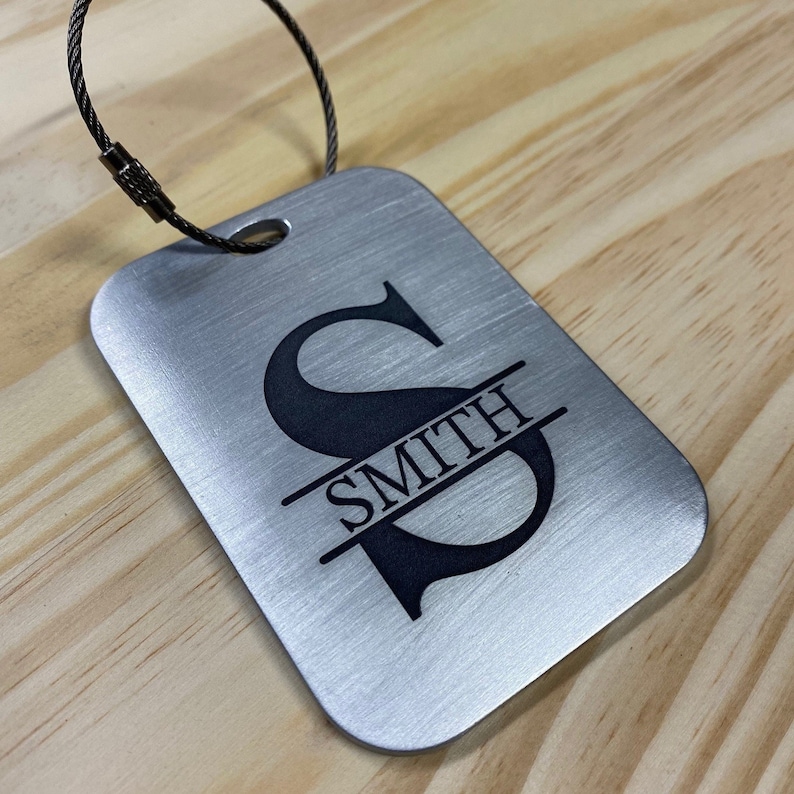 May include: Silver luggage tag with a black engraved initial "S" and the name "SMITH" split by a line through the letter.