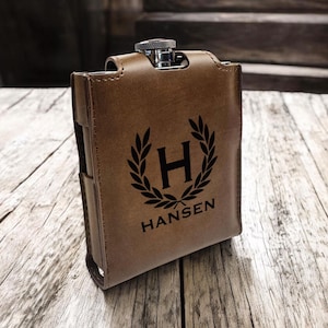 May include: A brown leather flask with a silver top and a personalised engraving. The engraving features a laurel wreath with the letter "H" in the centre and the name "Hansen" below.