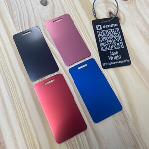 May include: Four metal luggage tags in different colors: black, rose gold, red, and blue. Each tag has a hole for a strap and a small hole at the top. The black tag has a QR code and text that reads "venmo Josh Wright @wrightswoodworks".