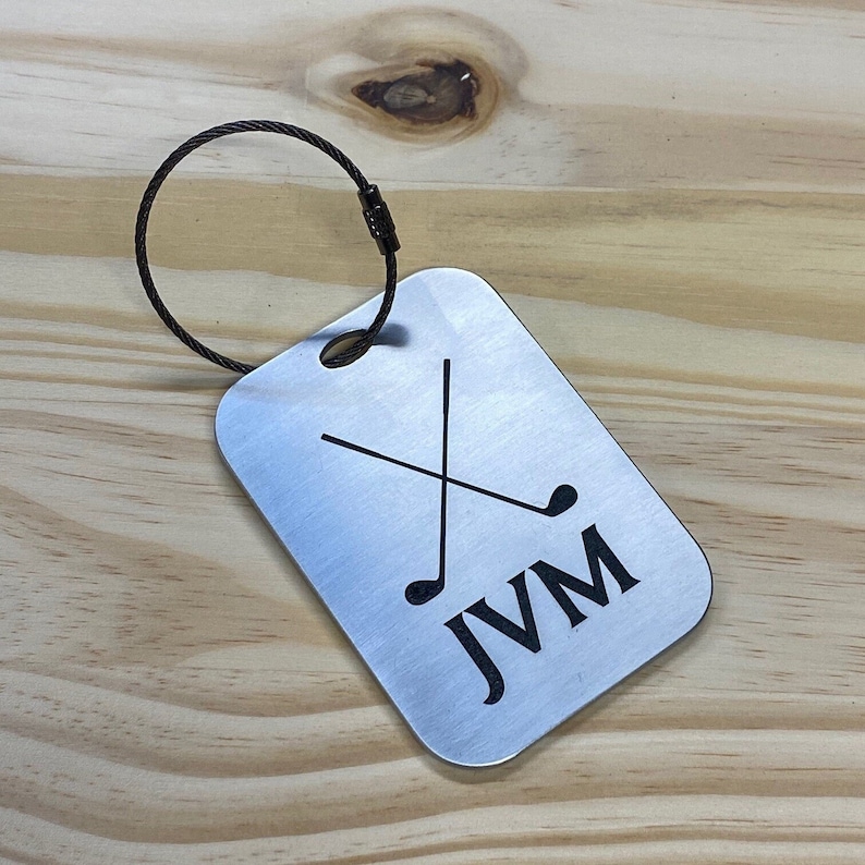 May include: Silver luggage tag with a black golf club design and the initials "JVM" engraved on it. The tag is attached to a black metal ring.