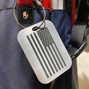 May include: A silver luggage tag with a black and white American flag design. The tag is attached to a silver chain with a black and white hand logo.