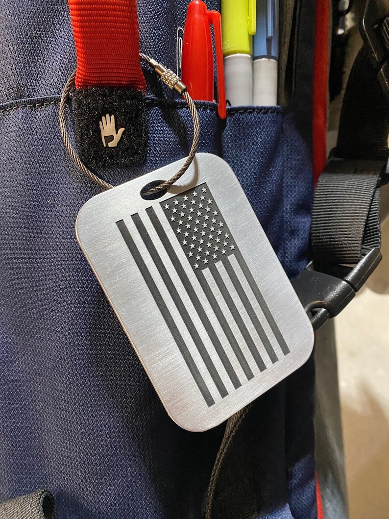 May include: A silver luggage tag with a black and white American flag design. The tag is attached to a silver chain with a black and white hand logo.