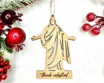 May include: Wooden Christmas ornament featuring a silhouette of Jesus with outstretched arms. The ornament is painted in a light brown color and has the text "Think celestial" engraved on the bottom.