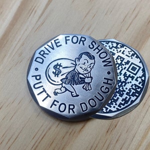 May include: Silver-toned metal golf ball marker with a black and white cartoon illustration of a person carrying a bag of money. The text "Drive for Show, Putt for Dough" is engraved around the edge of the marker. The back of the marker has a black and white QR code.