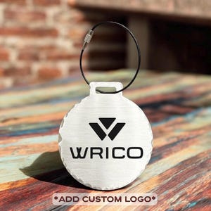 May include: A silver-colored, round keychain tag with a black logo and the word "WRICO" printed on it. The tag has a textured surface and a black wire loop for attachment. The bottom of the tag reads "*ADD CUSTOM LOGO*".