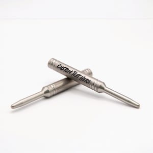 May include: Two metallic, silver-toned cylindrical tools with tapered ends and grooved detailing near the center. The top tool features the black printed text CUSTOM TEXT HERE along its barrel. Both items are positioned in a crossed arrangement.