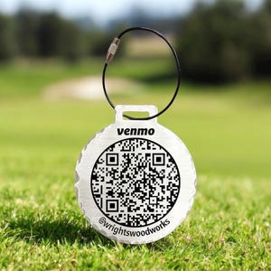 May include: A round, silver-colored tag with a black QR code and the word "venmo" printed on it. The tag is attached to a black metal loop. The text "@wrightswoodworks" is printed at the bottom of the tag. The tag is on a green grassy background.