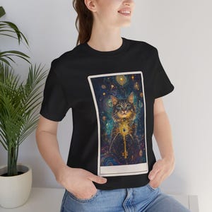 May include: Black t-shirt featuring a detailed illustration of a cat holding a golden key. The cat is set against a cosmic background with celestial elements. The design is framed within a white rectangle.