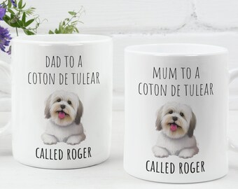 May include: Two white ceramic mugs with black text and a cartoon image of a white Coton de Tulear dog. The text on the mugs reads "Dad to a Coton de Tulear Called Roger" and "Mum to a Coton de Tulear Called Roger".