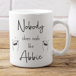 May include: White ceramic mug with black text that reads "Nobody does nails like Abbie". The text is surrounded by two black hand drawings with manicured nails.