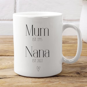 Puede incluir: Taza de cer&aacute;mica blanca con las palabras "Mum EST. 1995 Nana EST. 2022" en escritura negra, con un peque&ntilde;o s&iacute;mbolo de coraz&oacute;n. La taza est&aacute; sobre una superficie de madera, con un jarr&oacute;n blanco de margaritas al fondo.