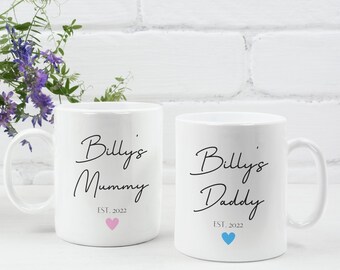 May include: Two white ceramic mugs with black text. The mug on the left says "Billy's Mummy Est. 2022" with a pink heart. The mug on the right says "Billy's Daddy Est. 2022" with a blue heart.