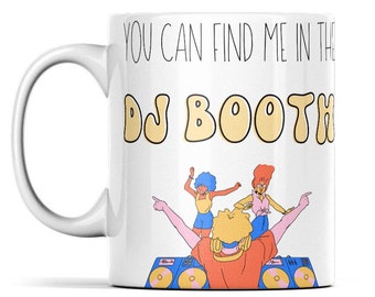 May include: White ceramic mug with a black outline and the text "You can find me in the DJ Booth". The text is in a yellow bubble font. There is a cartoon illustration of a DJ with headphones and two people dancing in front of the DJ.