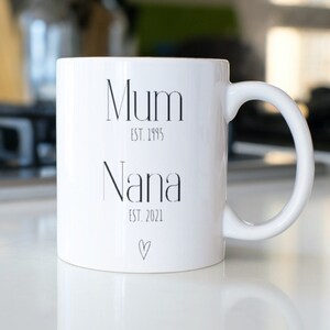 Puede incluir: Taza de cer&aacute;mica blanca con las palabras "Mum" y "Nana" impresas en negro, junto con los a&ntilde;os "EST. 1995" y "EST. 2021". Un peque&ntilde;o coraz&oacute;n est&aacute; impreso debajo de "Nana". La taza tiene un asa curva.