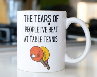 May include: White ceramic mug with black text that reads "The Tears of People I've Beat at Table Tennis" and a drawing of two ping pong paddles.