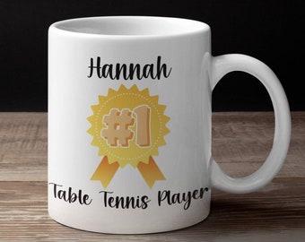 May include: White ceramic mug with a black design that says "Hannah #1 Table Tennis Player" with a gold medal graphic.