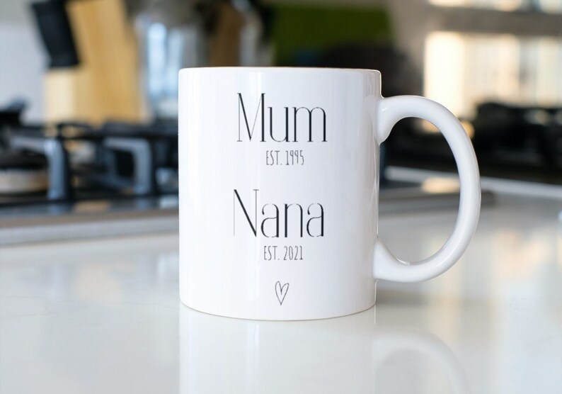 Puede incluir: Taza de cer&aacute;mica blanca con las palabras "Mum" y "Nana" impresas en negro, junto con los a&ntilde;os "EST. 1995" y "EST. 2021". Un peque&ntilde;o coraz&oacute;n est&aacute; impreso debajo de "Nana". La taza tiene un asa curva.