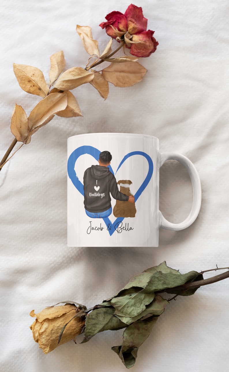 May include: White ceramic mug with a blue heart design. The heart features a cartoon illustration of a person with a dog. The mug has the text "I love Bulldogs" and "Jacob & Bella" printed on it.