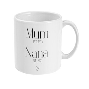 Puede incluir: Taza de cer&aacute;mica blanca con las palabras "Mum EST. 1995" y "Nana EST. 2021" impresas en negro, junto con un peque&ntilde;o s&iacute;mbolo de coraz&oacute;n. La taza tiene un asa curvada y un dise&ntilde;o sencillo.