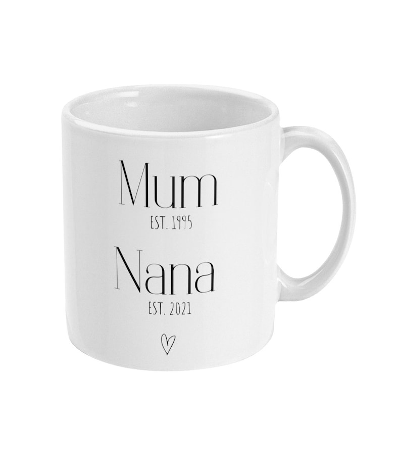 Puede incluir: Taza de cer&aacute;mica blanca con las palabras "Mum EST. 1995" y "Nana EST. 2021" impresas en negro, junto con un peque&ntilde;o s&iacute;mbolo de coraz&oacute;n. La taza tiene un asa curvada y un dise&ntilde;o sencillo.