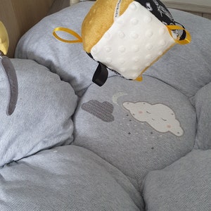 May include: A baby play mat with a hanging toy. The mat is light grey with a striped pattern and cloud and moon designs. The hanging toy is a cube with yellow, white, and black sides, and a small yellow ribbon.