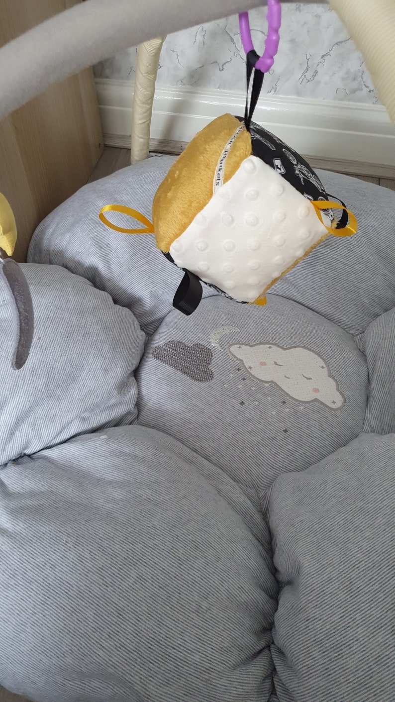 May include: A baby play mat with a hanging toy. The mat is light grey with a striped pattern and cloud and moon designs. The hanging toy is a cube with yellow, white, and black sides, and a small yellow ribbon.