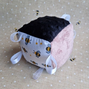 May include: A tactile baby cube in black, pink, and patterned fabrics. The cube has a black plush top, a pink plush side, and a white fabric side decorated with yellow polka dots and bee illustrations. White ribbon loops are attached for sensory play.
