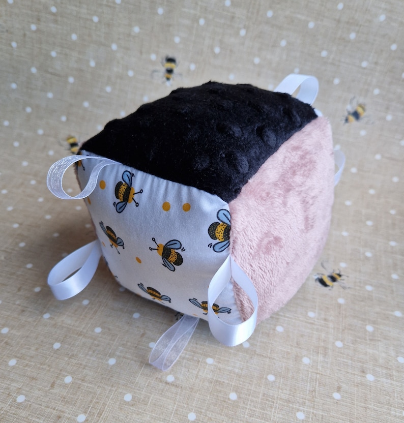 May include: A tactile baby cube in black, pink, and patterned fabrics. The cube has a black plush top, a pink plush side, and a white fabric side decorated with yellow polka dots and bee illustrations. White ribbon loops are attached for sensory play.