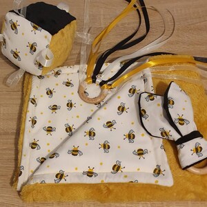 May include: A baby sensory set with a bee theme in yellow and white. The set includes a cube, a blanket, and a teether. The cube has black and yellow sides with white ribbons. The blanket and teether have a bee pattern and wooden rings.