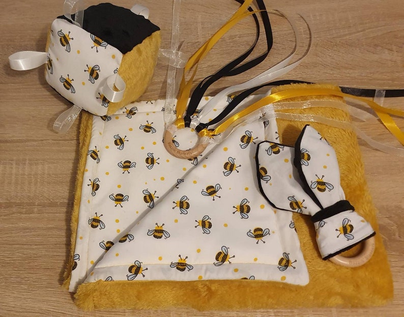 May include: A baby sensory set with a bee theme in yellow and white. The set includes a cube, a blanket, and a teether. The cube has black and yellow sides with white ribbons. The blanket and teether have a bee pattern and wooden rings.