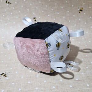 May include: A soft baby block in black, pink, and white. The white side has a yellow bee pattern. White satin ribbons are attached to the block. A baby toy, designed for sensory play.