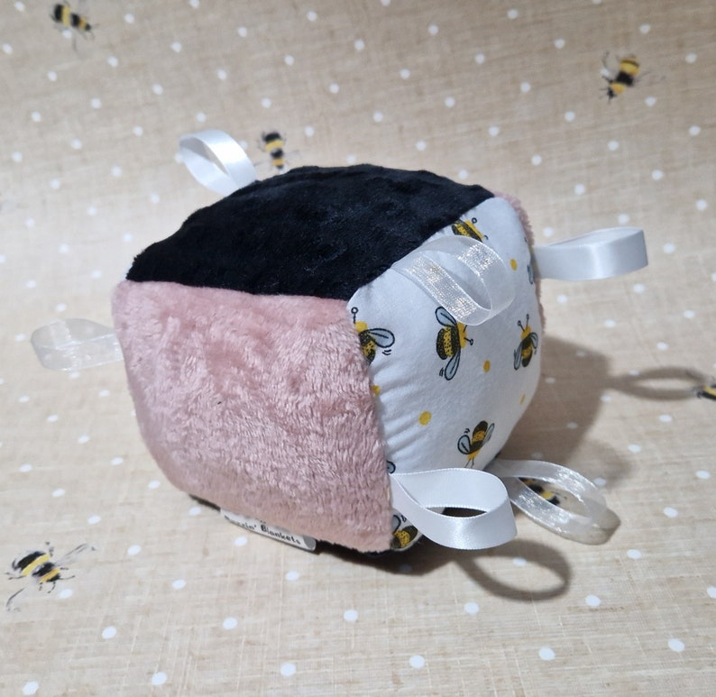 May include: A soft baby block in black, pink, and white. The white side has a yellow bee pattern. White satin ribbons are attached to the block. A baby toy, designed for sensory play.