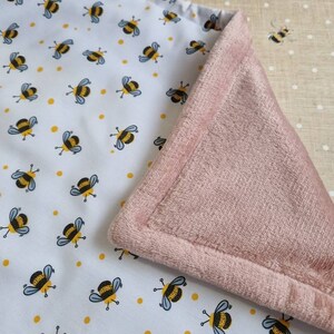 May include: A baby blanket with a white top layer patterned with cartoon bees and yellow dots. The underside is a soft, pink plush material. The blanket is folded to show both sides.