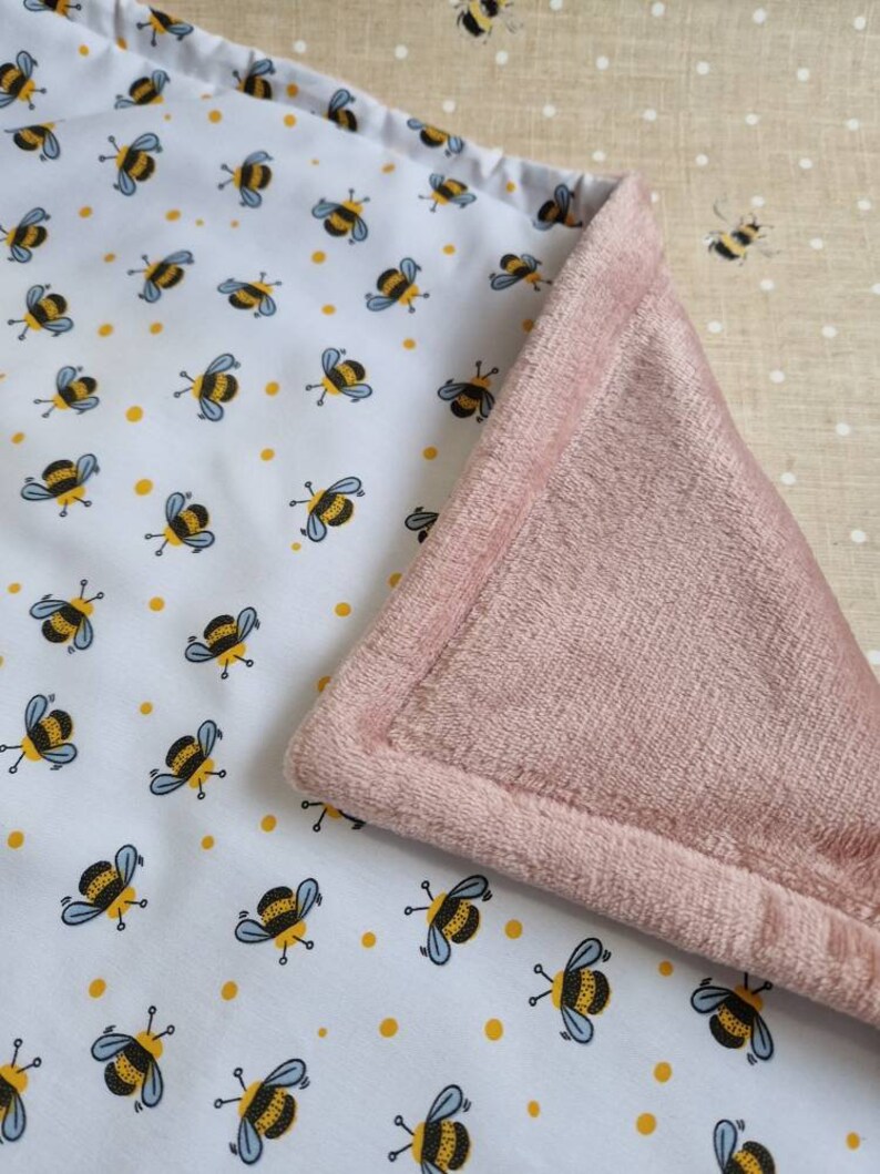 May include: A baby blanket with a white top layer patterned with cartoon bees and yellow dots. The underside is a soft, pink plush material. The blanket is folded to show both sides.