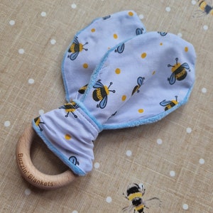 May include: A baby rattle featuring a wooden ring and soft, light blue fabric shaped like bunny ears. The fabric is patterned with yellow spots and cartoon bees. The wooden ring is engraved with "Buzzin' Blankets".