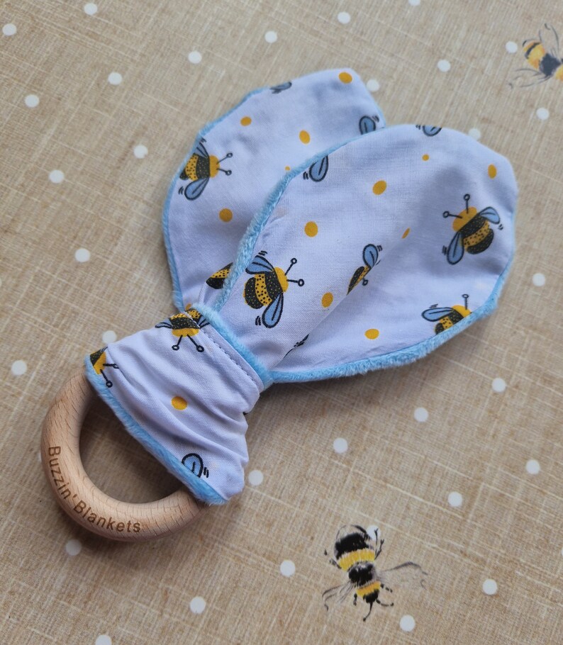May include: A baby rattle featuring a wooden ring and soft, light blue fabric shaped like bunny ears. The fabric is patterned with yellow spots and cartoon bees. The wooden ring is engraved with "Buzzin' Blankets".