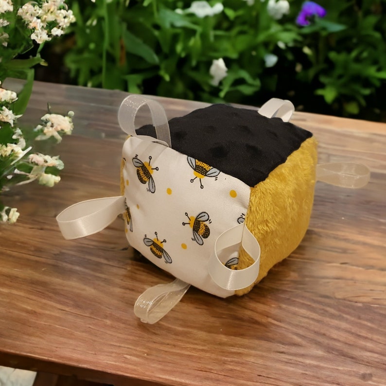 May include: A tactile baby block with a bee design. The cube has a black, plush top, a yellow side, and a white fabric side decorated with yellow polka dots and black and yellow bee illustrations. White ribbon loops are attached to each side.