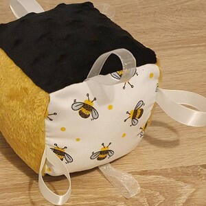 May include: A tactile cube toy with a bee design. The cube has panels of black, yellow, and white fabric. One side displays a bee pattern, another is black, and a third is yellow. White ribbons are attached for sensory play.