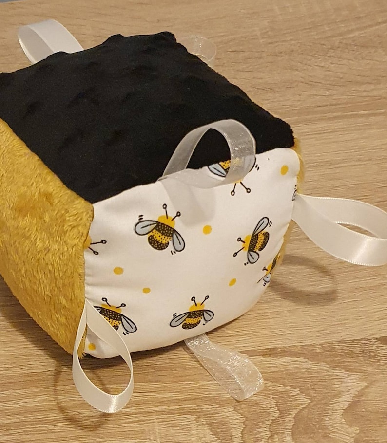 May include: A tactile cube toy with a bee design. The cube has panels of black, yellow, and white fabric. One side displays a bee pattern, another is black, and a third is yellow. White ribbons are attached for sensory play.