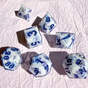 Peut inclure: Un ensemble de sept dés polyédriques en résine blanche avec un motif floral et d'insectes bleu incrusté. Chaque dé présente des chiffres bleu métallique gravés sur les faces, incluant un d20, d12, d10, d8, d6, d4 et un dé de pourcentage.