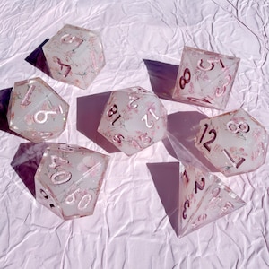 May include: A set of seven polyhedral dice made of translucent off-white resin with embedded pink floral patterns and glitter. Each die features engraved numbers filled with metallic pink paint, including a d20, d12, d10, d8, d6, d4, and percentile die.