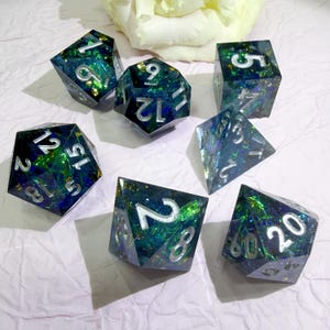 May include: A set of seven polyhedral dice featuring a dark blue and green translucent resin base filled with shimmering iridescent flakes. Each die has deeply engraved numbers painted in a bright silver finish for high contrast and readability.